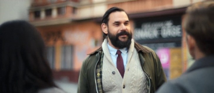 Outdoor scene from the German Netflix movie Brick. Medium closeup shot of a bearded white guy with a shirt and tie who is standing in the street facing the camera. Two other actors, a brunette woman on the left and a man on the right are with their backs to the camera. Due to the shallow depth of field and motion blur even the actor who should be in focus is slightly out of focus. The street background is very out of focus as are the backs of the other to actors.