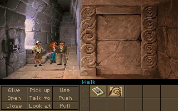 In-game screenshot of Indiana Jones and the Fate of Atlantis. It shows the inside of a temple with beige stone walls and stone floor in a pixelized art style. Three people are standing in the room (Indiana Jones in brown leather clothing, a woman with long red hair wearing blue trousers and another man wearing a tropical explorer outfit).
The bottom quarter of the screen is filled with activity verbs like give, open, push or pull as well as items the player has collected. A magazine and Indy's…