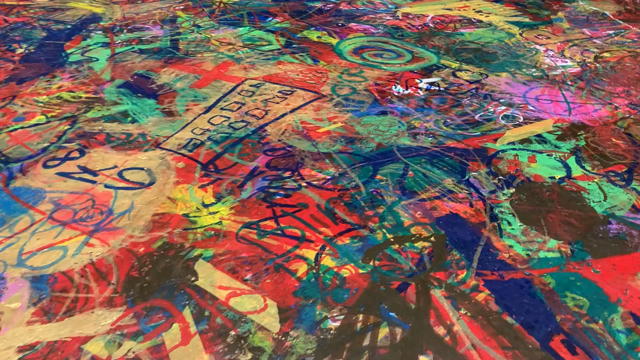 The camera moves closely along a floor that is covered by multiple layers of colourful and random scribbles. They have been made by visitors to the museum using wax crayons. At the end of the video the camera pans up to reveal a view of the whole room. A tall attium, ca 20 by 40 meters, with a brown marble floor (all covered in crayon scribbles), white columns and overhead lighting. A small number of visitors is strolling across the room.