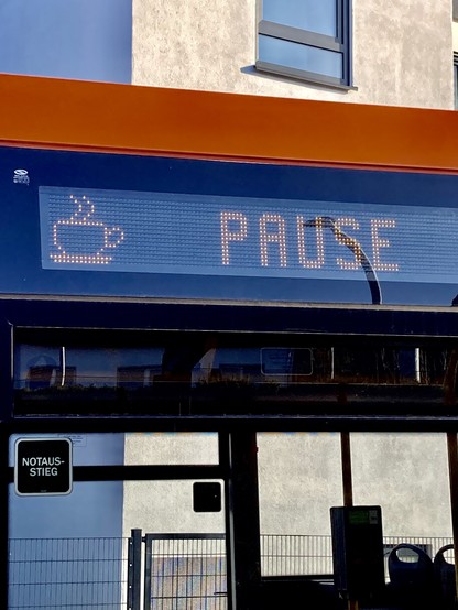 An LED sign on the side of a bus displays the word pause and a pixelated steaming cup of coffee. 
The photo itself has strong horizontal and vertical lines made up by the large window frame and the building behind it.