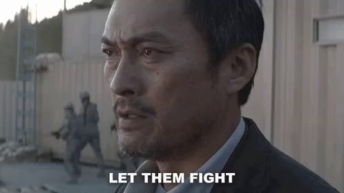 Japanese actor Ken Watanabe, a determined-looking man in a suit, is saying: "let them fight" while soldiers are running around in the background.